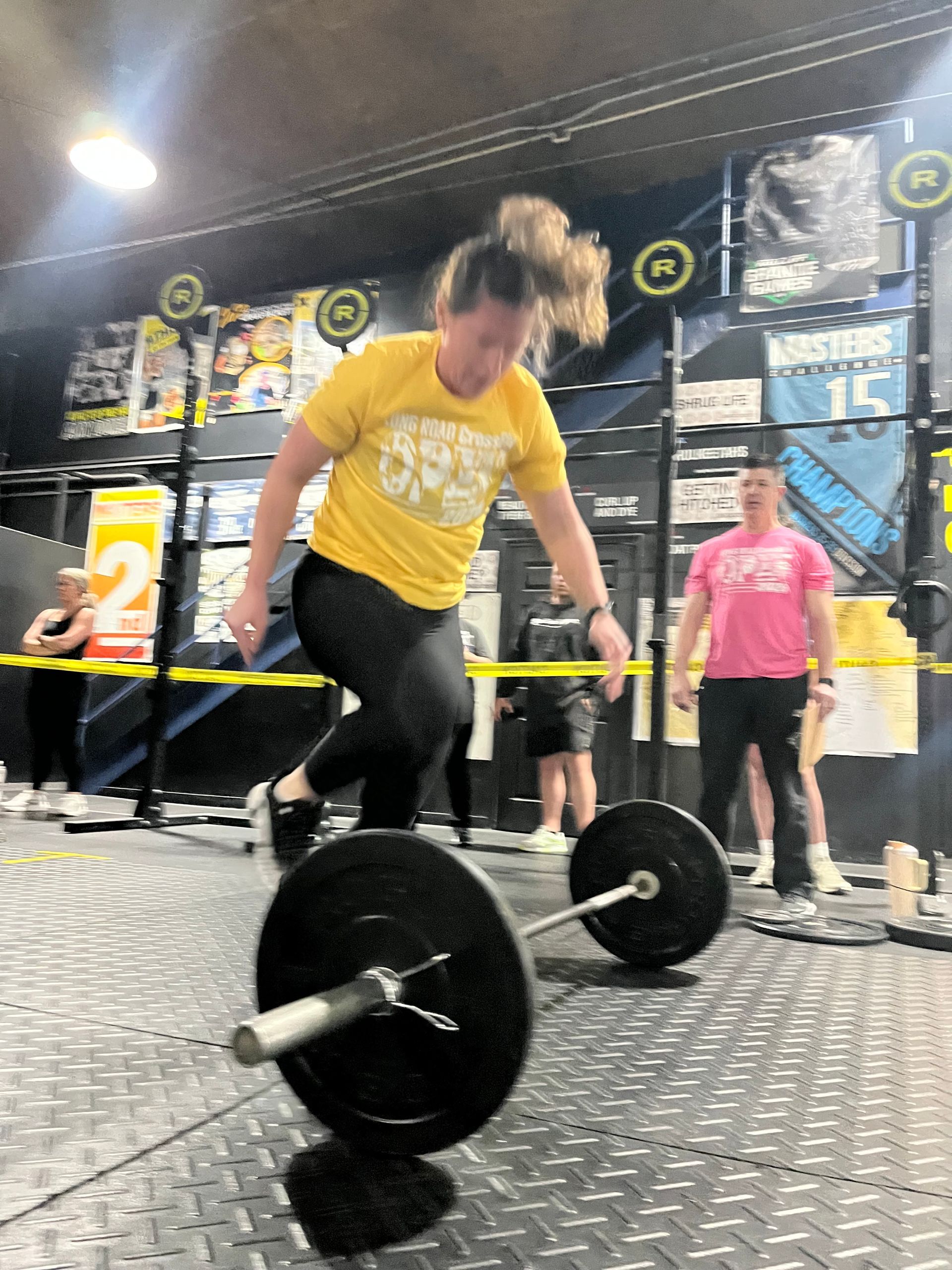 Person in yellow shirt lifting a barbell on a gym floor with spectators in the background