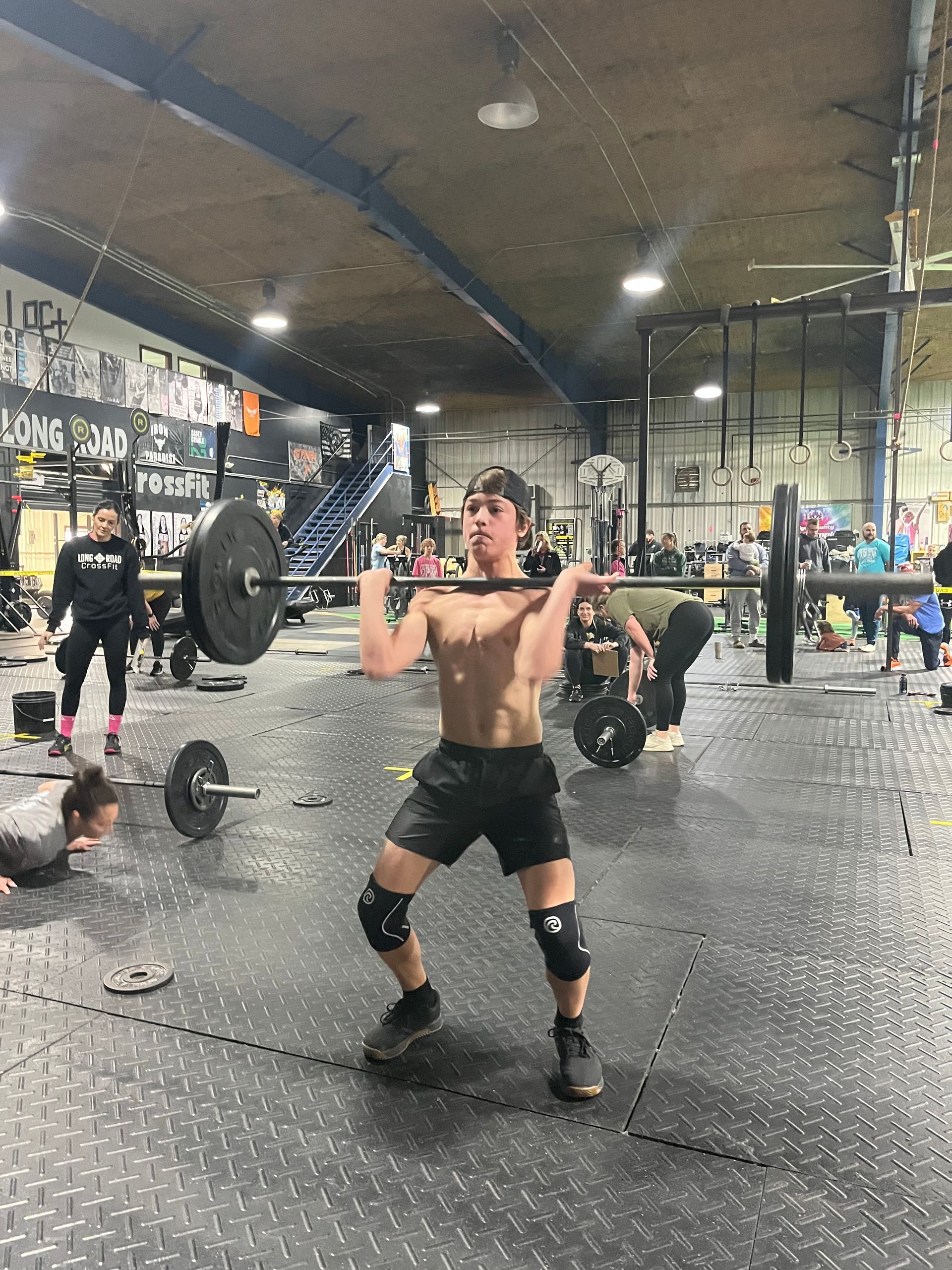 Man lifting a barbell overhead in a CrossFit gym, with other athletes and equipment in the background