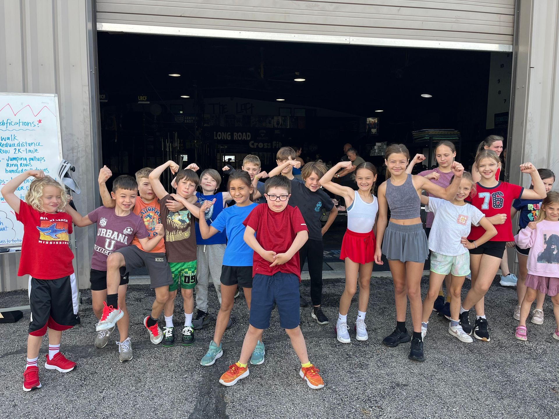 Group of children posing and flexing outside a garage, smiling in colorful athletic clothes.