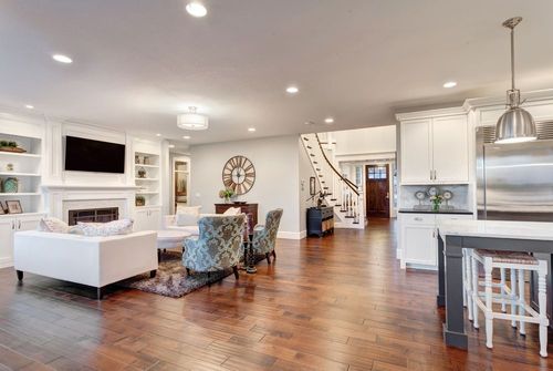 Bright open-plan living room and kitchen with white furniture, hardwood floors, and stairs in the background