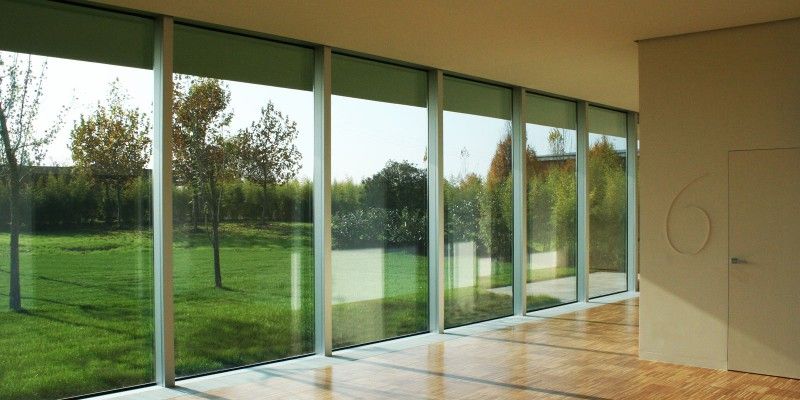 Camera moderna con pareti vetrate a tutta altezza che si affacciano su un prato verde e alberi, inondata di luce solare.
