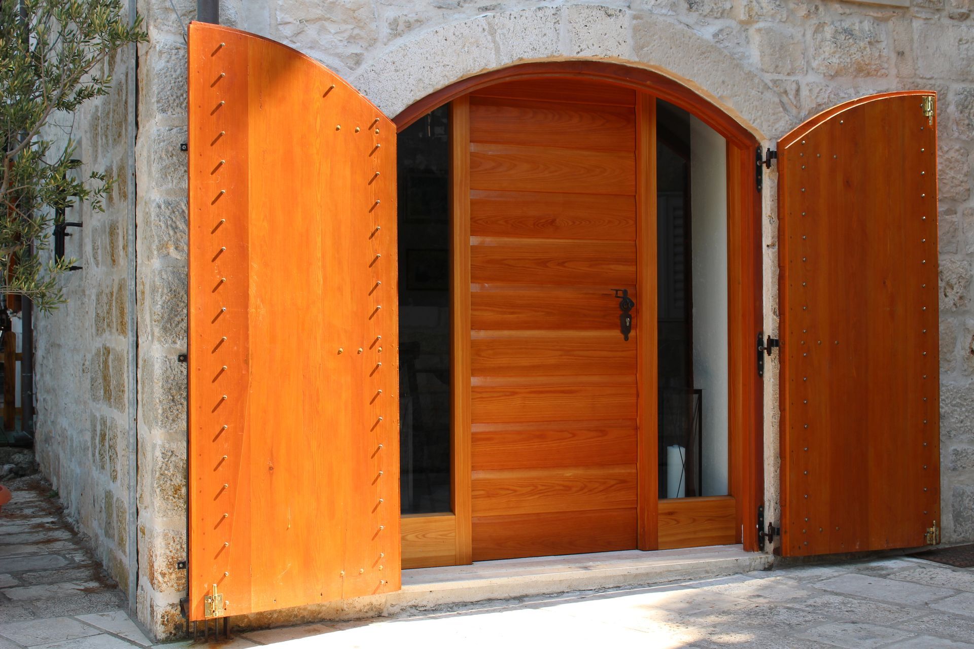 Doppie porte di legno aperte in un arco di pietra, con la luce del sole che entra dall'alto.