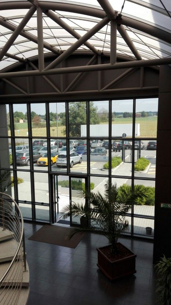 Atrio interno con ampia parete vetrata affacciata su un parcheggio e sull'acqua, con travi in acciaio a vista nella parte superiore.