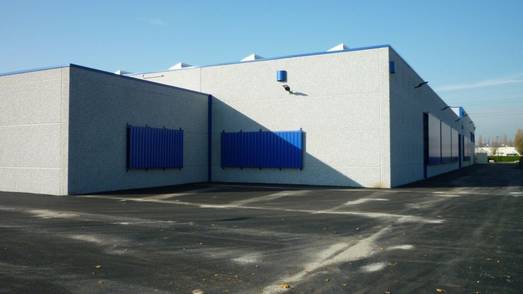 Esterno di un magazzino industriale con pareti bianche, porte blu e piazzale pavimentato esposto al sole.