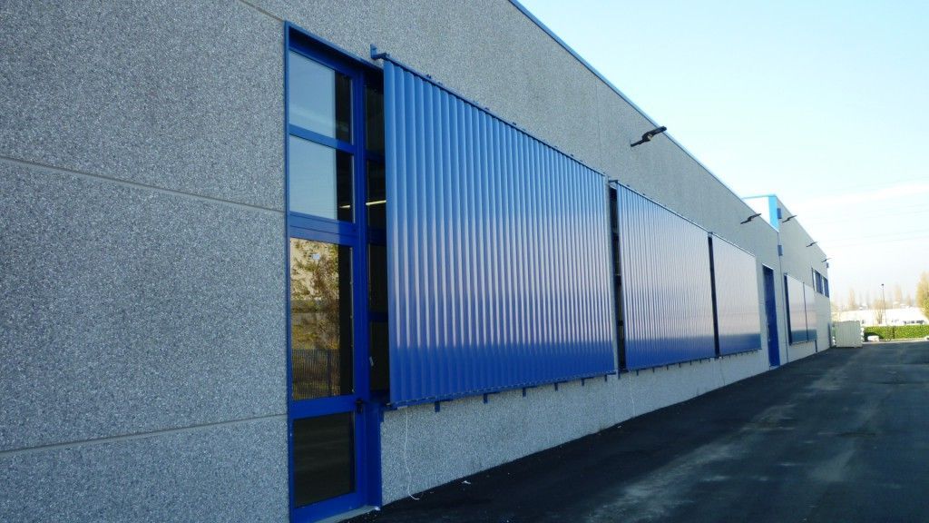 Edificio industriale blu con rivestimento in lamiera grecata e finestre strette, adiacente a un lotto pavimentato vuoto.
