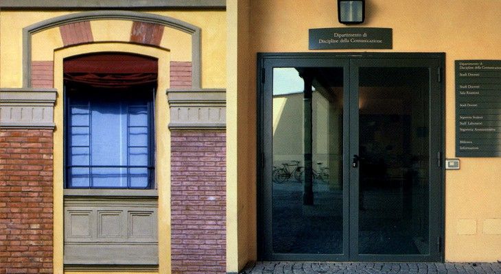 Ingresso dell'edificio con porte in vetro scuro, facciata con finestre blu e insegne a muro sopra e accanto alla porta.