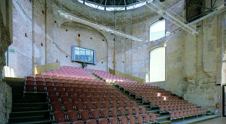 Auditorium abbandonato con file di sedili rossi e un soffitto danneggiato sotto una grande cupola.