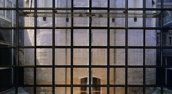Facciata a griglia in vetro e acciaio con un portale illuminato da una luce calda al centro.