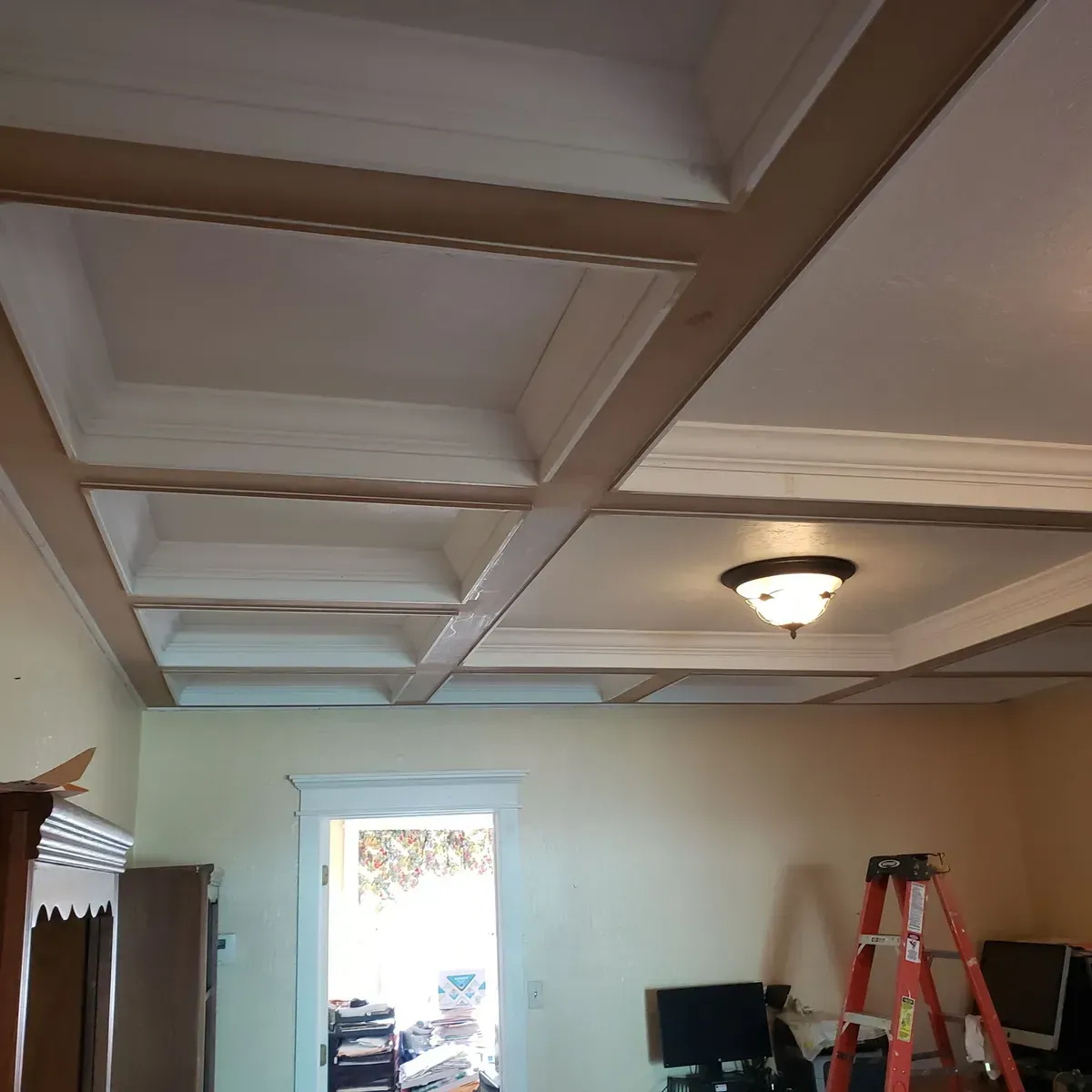 Coffered ceiling in a room with a central light, doorway, and a ladder near two monitors.