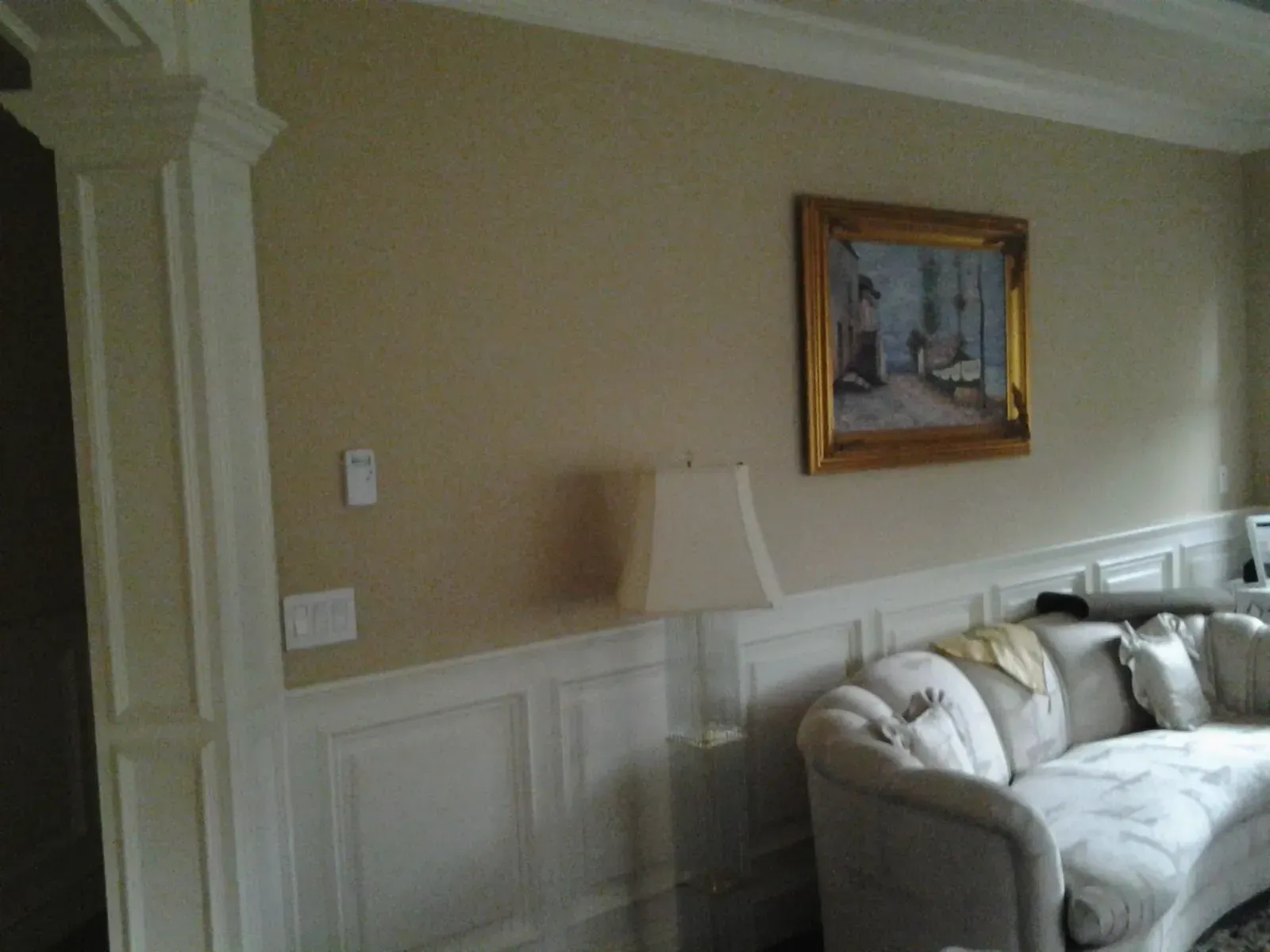Living room with beige walls, white wainscoting, a cream sofa, lamp, and framed painting on the wall
