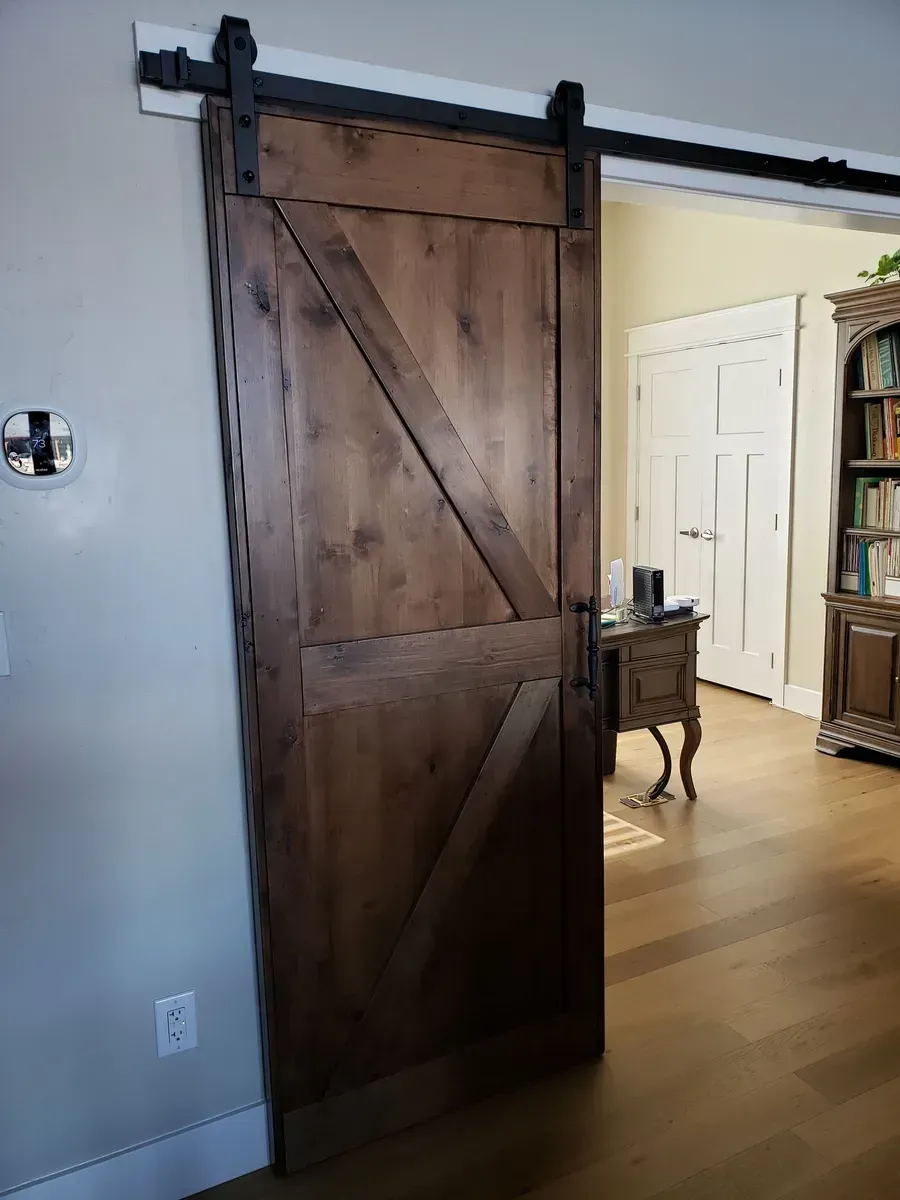 Wooden sliding barn door partially open to a bright hallway and bookshelf inside a home