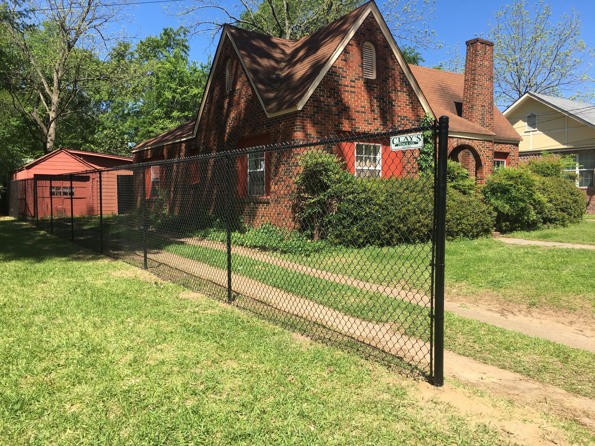 Black chain-link fence in front of a red brick house with a brown roof and a small shed on a sunny day.