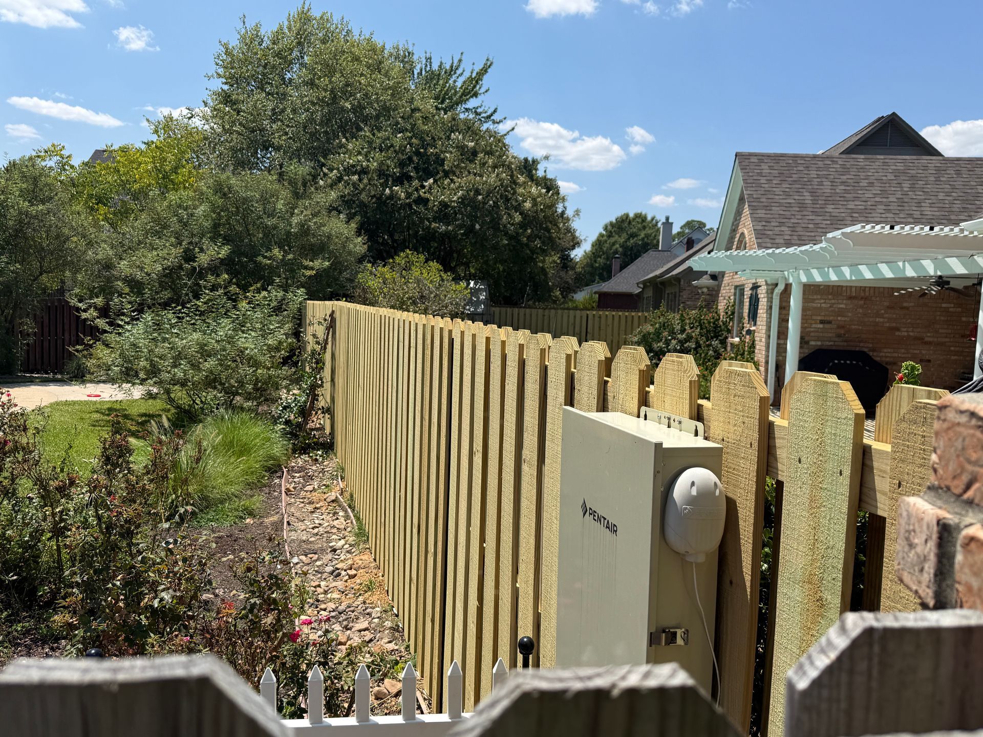 Wooden fence and gate beside a garden path, with trees, flowers, and a brick house under a blue sky.