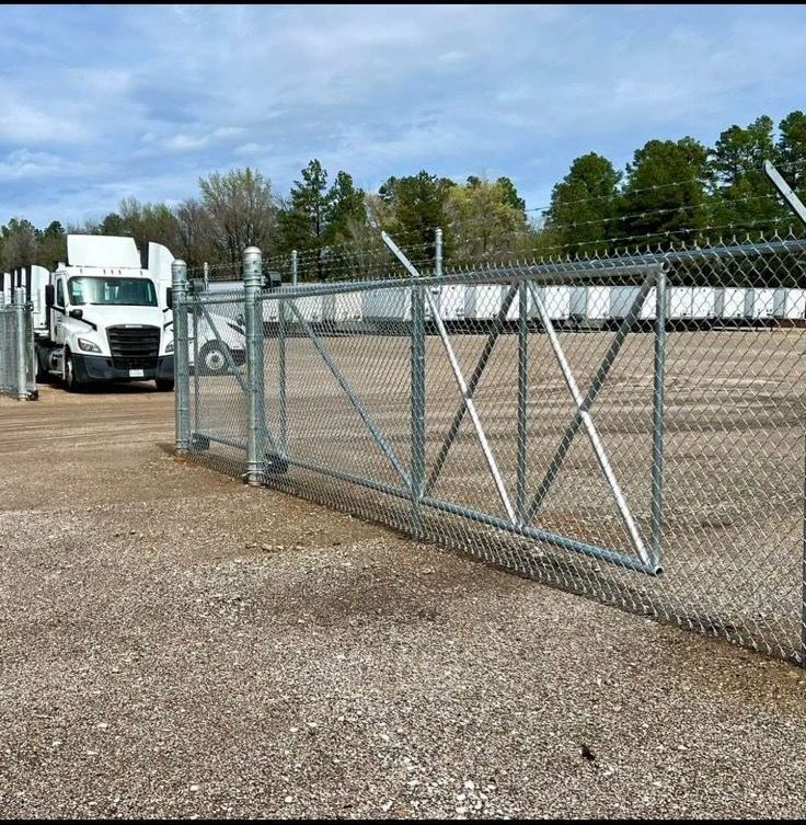 A chain link fence with barbed wire tops and a gate. A semi-truck approaches the open gate.