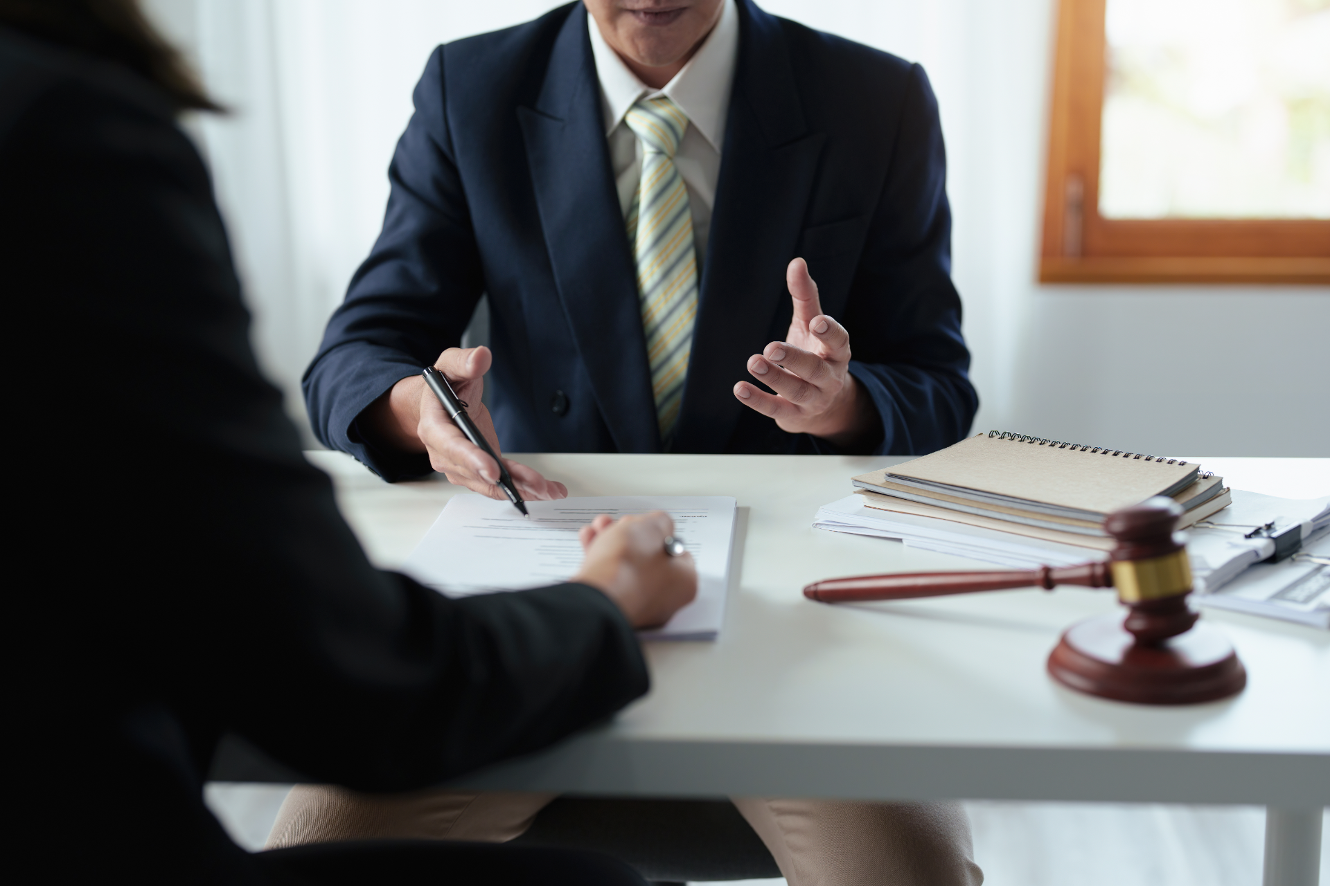 Lawyer in suit gestures, explaining paperwork to a person writing at a desk, gavel nearby.