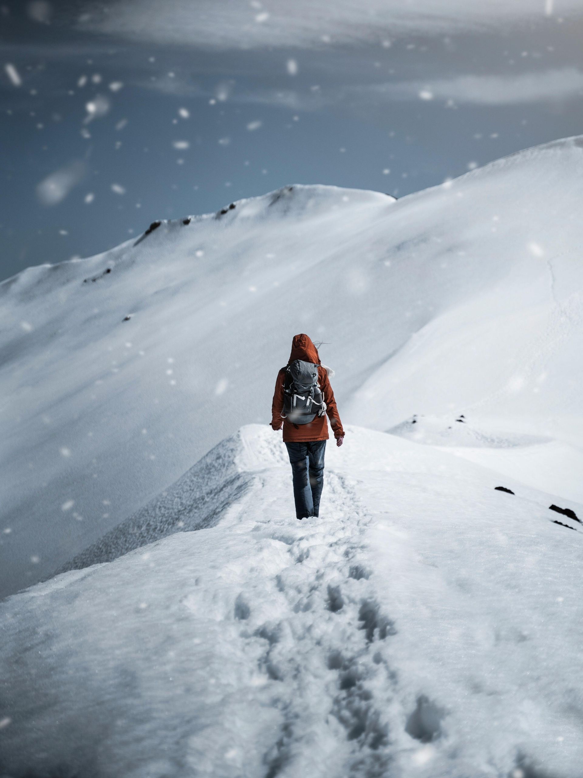 Lone hiker in an orange jacket walking along a snowy mountain ridge in falling snow