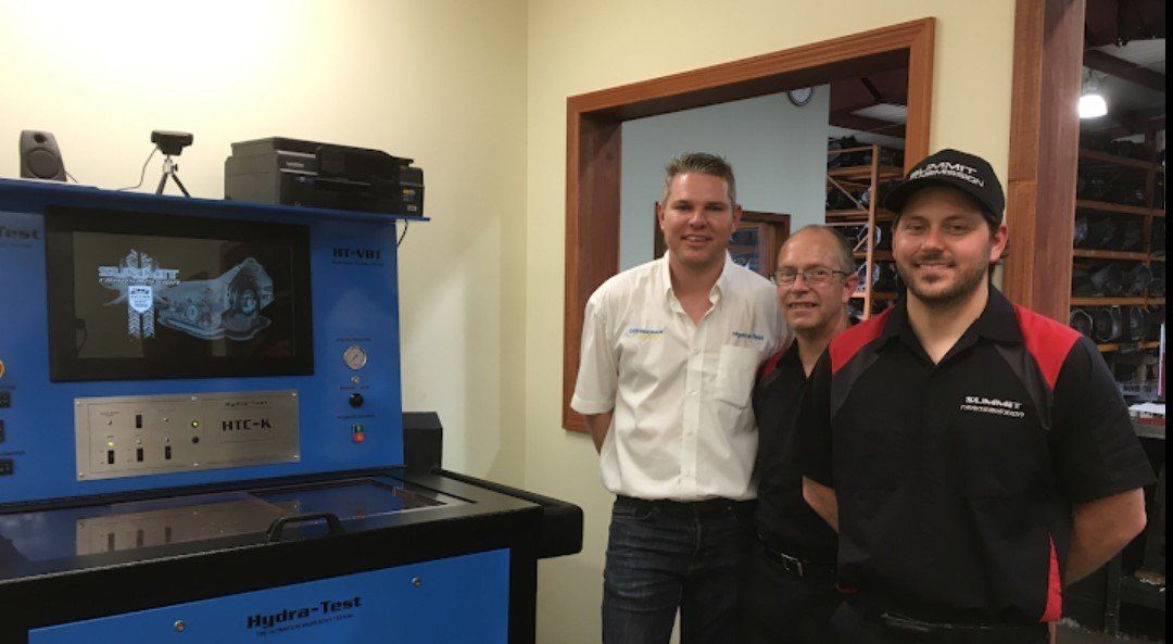 Three men stand by a blue testing machine. The machine displays an image. Inside a workshop.