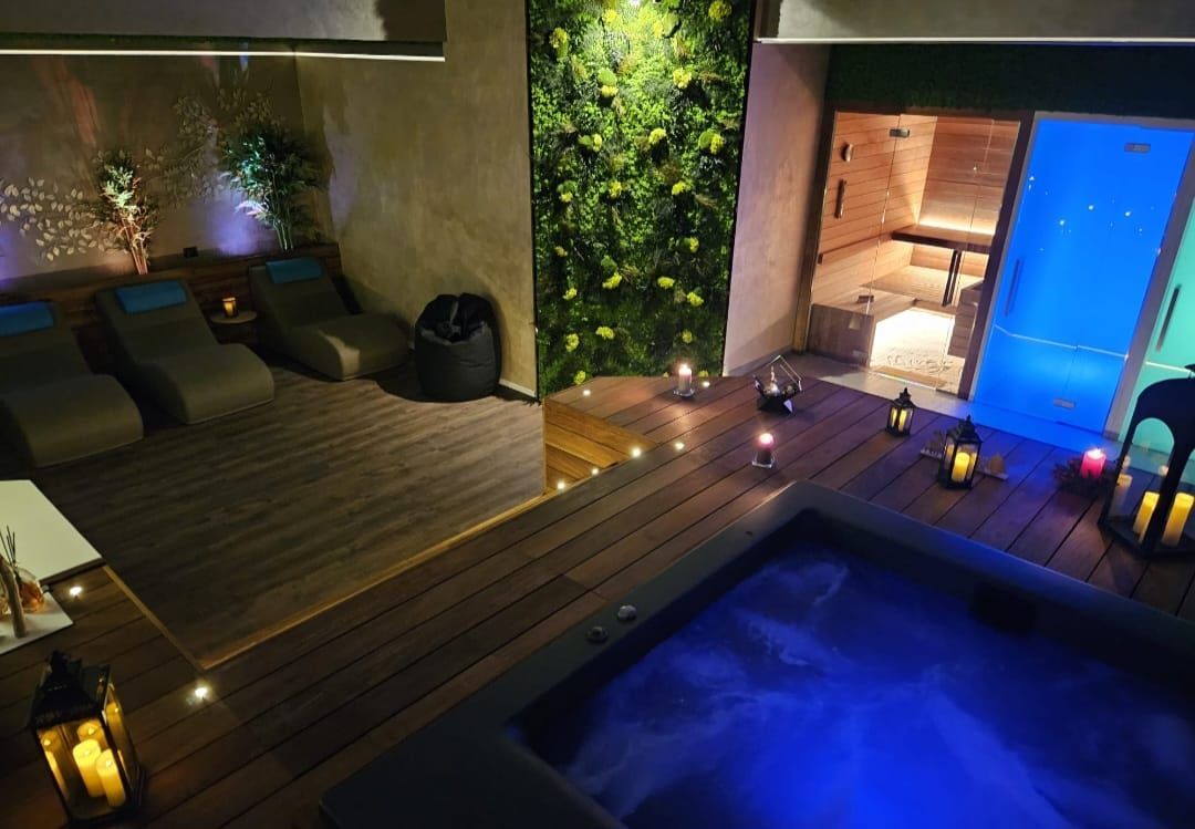 Area benessere con jacuzzi, terrazza in legno, sedie a sdraio, sauna e parete con giardino verticale. Candele.