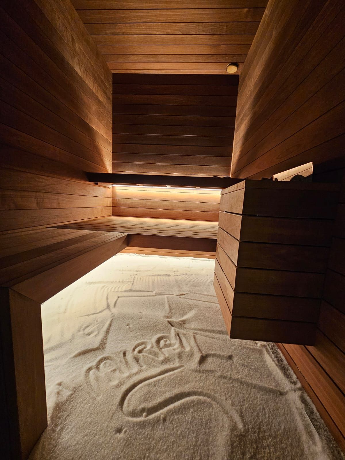 Interno della sauna in legno con pavimento in sabbia. Scritta sulla sabbia. Luci soffuse e panche in legno.