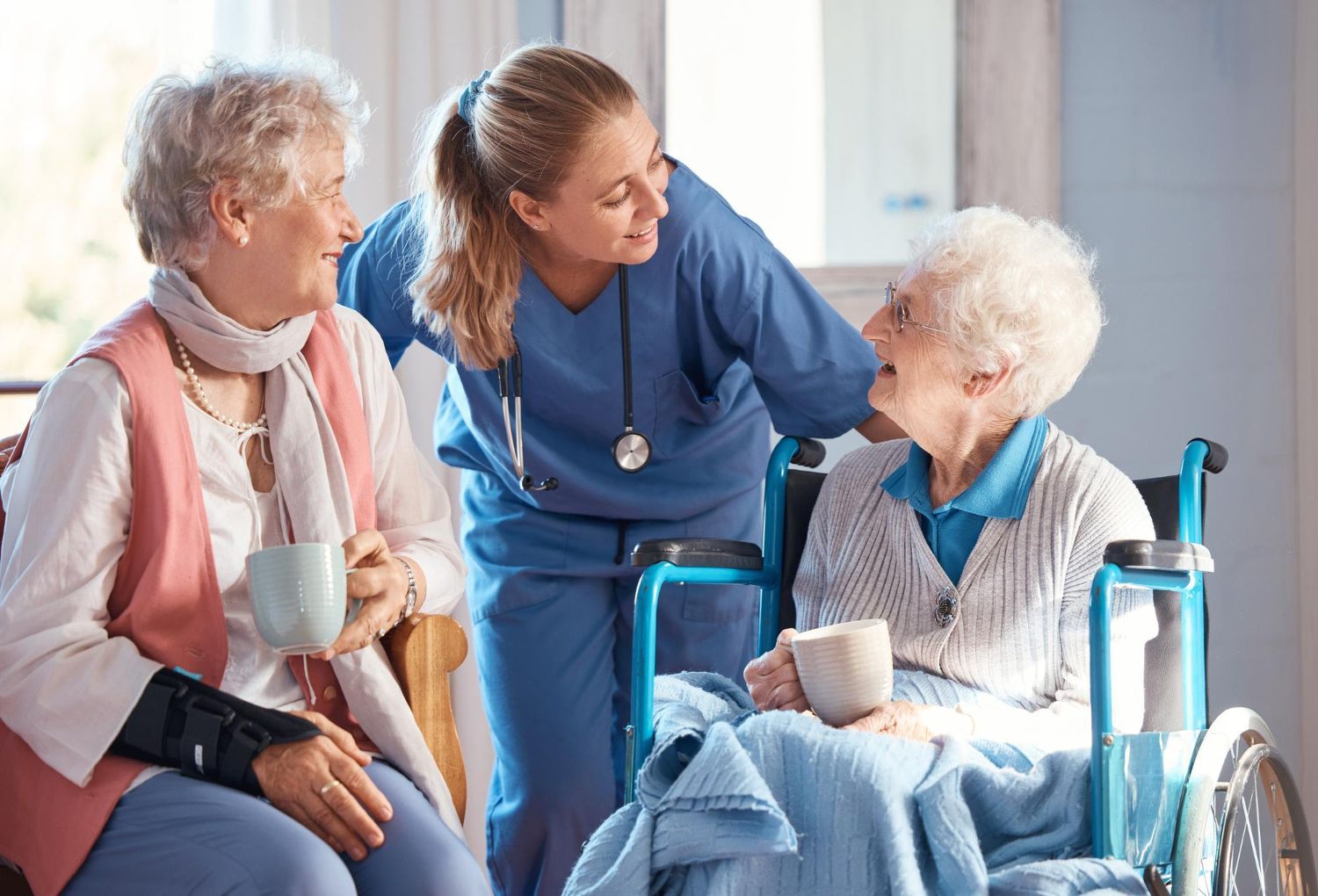 Eine Krankenschwester in blauer OP-Kleidung lächelt, während sie sich mit zwei Personen in einer Pflegeeinrichtung unterhält, die Tassen in der Hand halten und sich unterhalten.