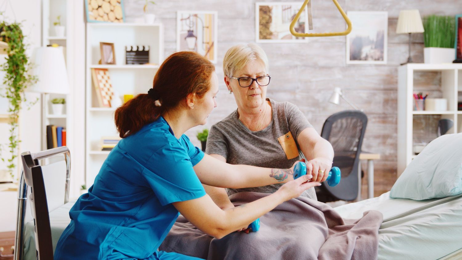 Eine medizinische Fachkraft in blauer OP-Kleidung hilft einem Patienten im Bett bei Armübungen mit kleinen Hanteln.