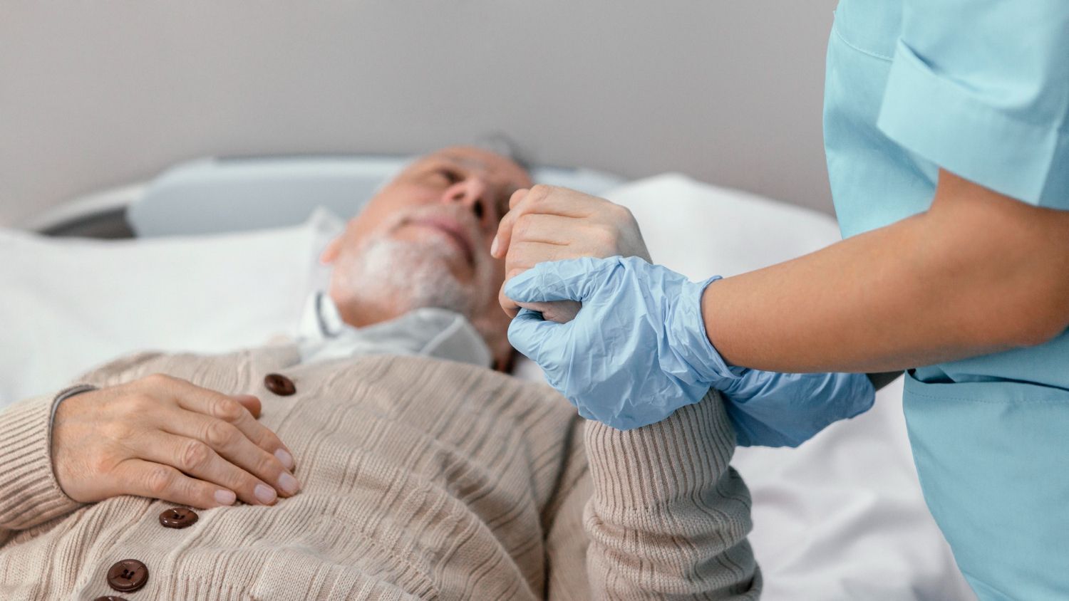 Eine in blauer OP-Kleidung und Handschuhen gekleidete Pflegekraft hält die Hand eines Patienten, der in einem Krankenhausbett ruht.