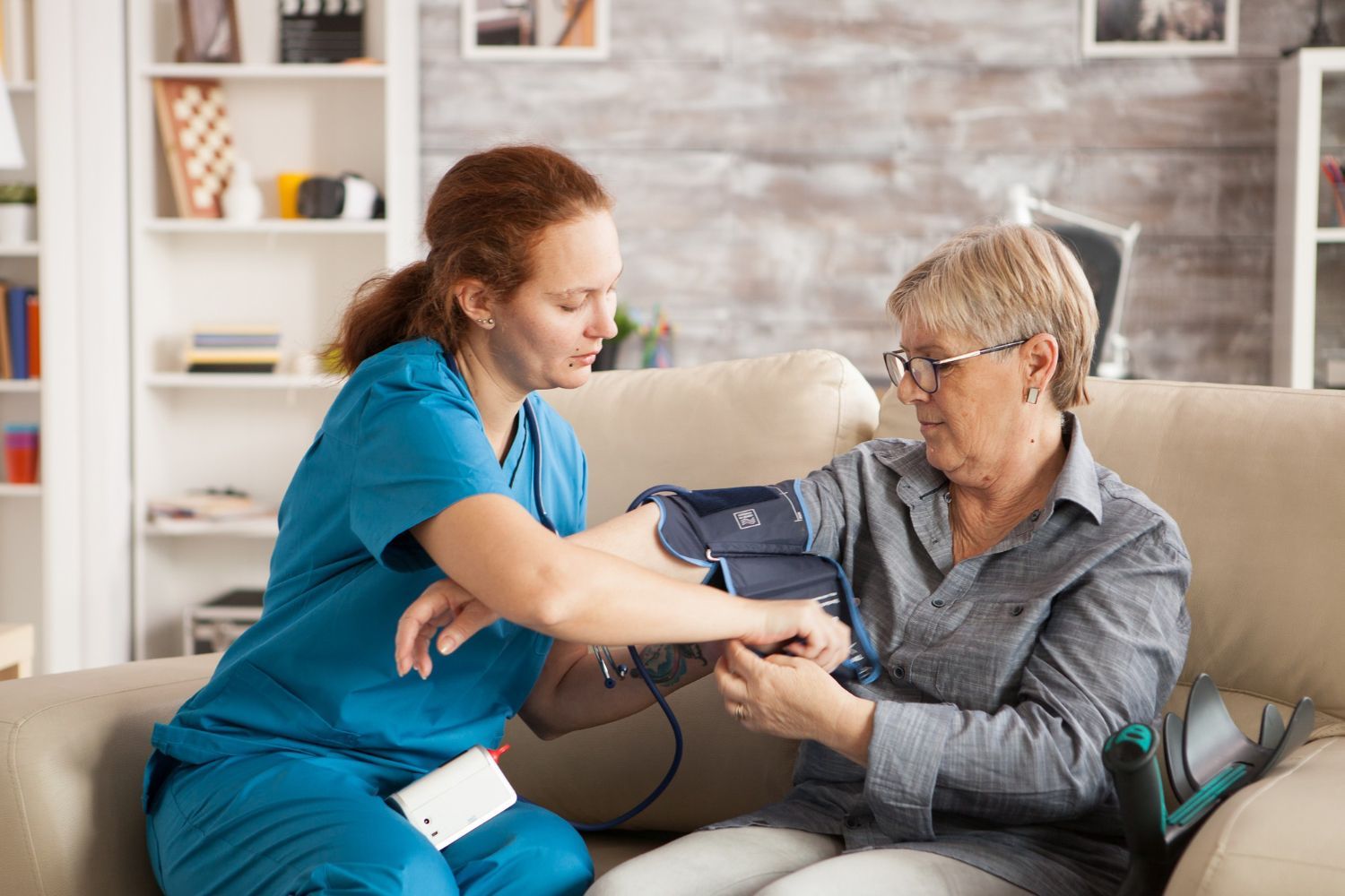 Eine medizinische Fachkraft in blauer OP-Kleidung misst den Blutdruck eines Patienten mit einer Blutdruckmanschette, während sie auf einer Couch in einem Haus sitzt.