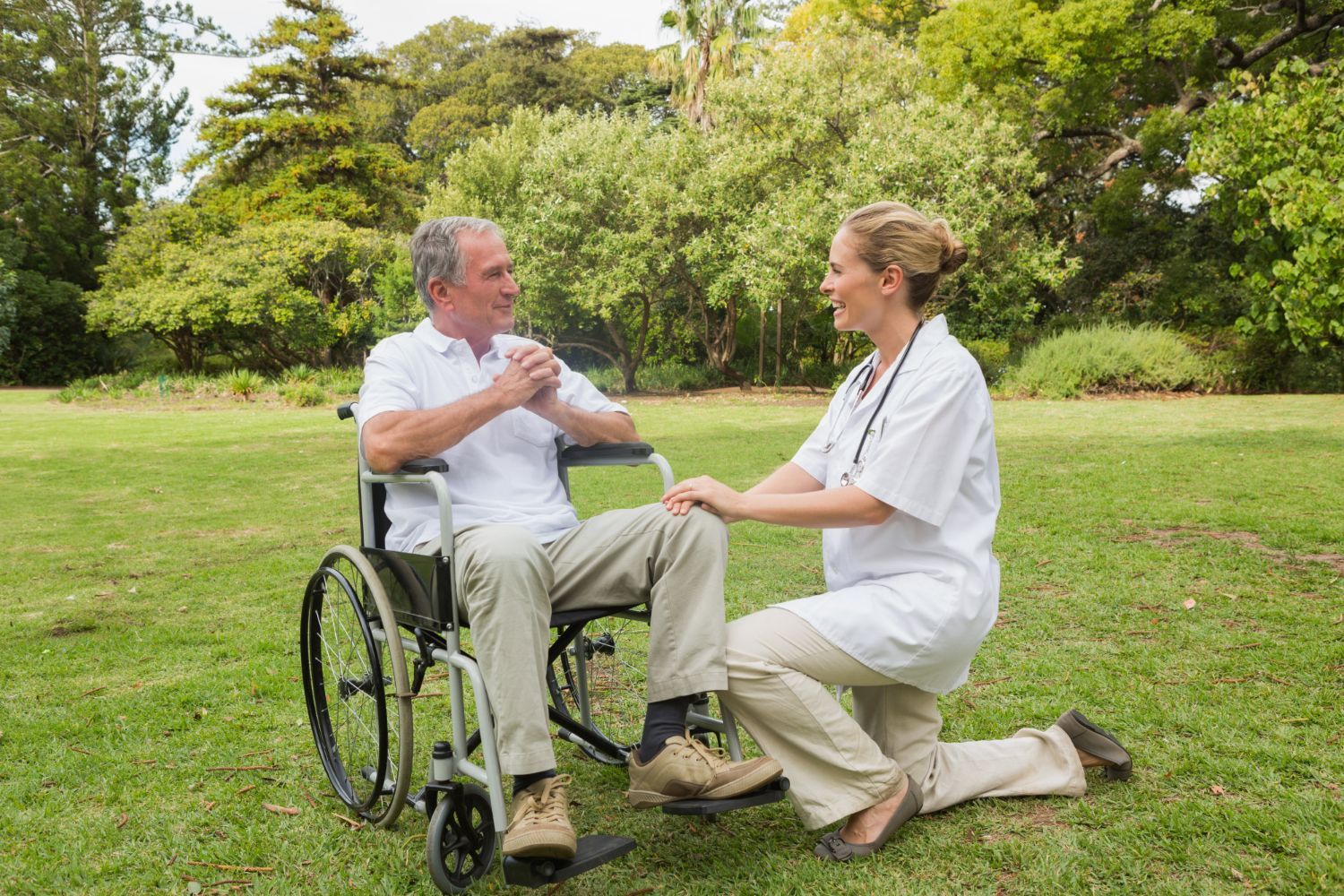 Eine Mitarbeiterin des Gesundheitswesens kniet neben einer Person im Rollstuhl, hält deren Hand und unterhält sich mit ihr in einem Park.
