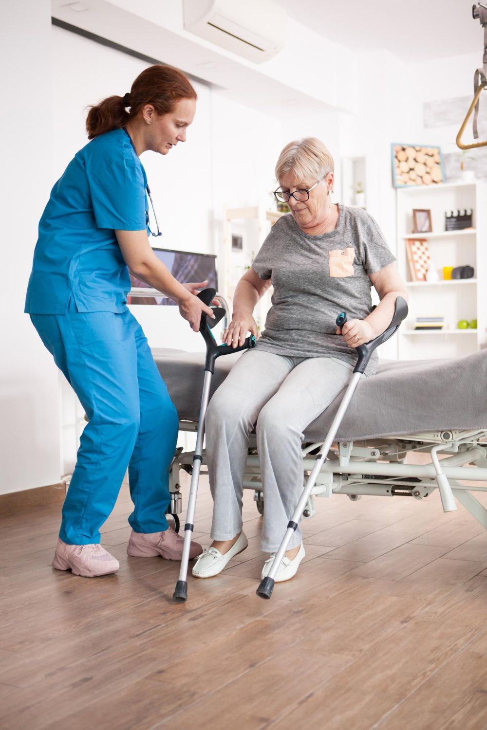 Eine medizinische Fachkraft in blauer OP-Kleidung hilft einem Patienten, der Unterarmgehstützen benutzt, vom Krankenhausbett aufzustehen.