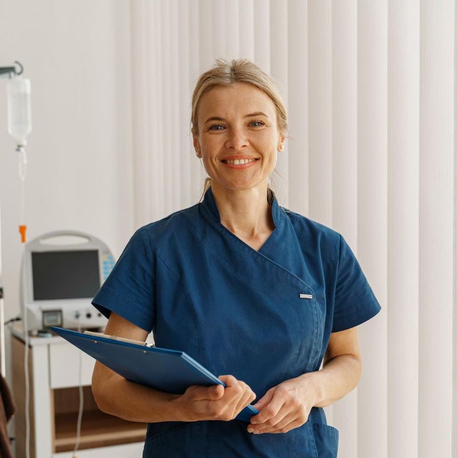 Eine lächelnde medizinische Fachkraft in blauer OP-Kleidung hält ein Klemmbrett in einer Klinik; im Hintergrund sind medizinische Geräte zu sehen.