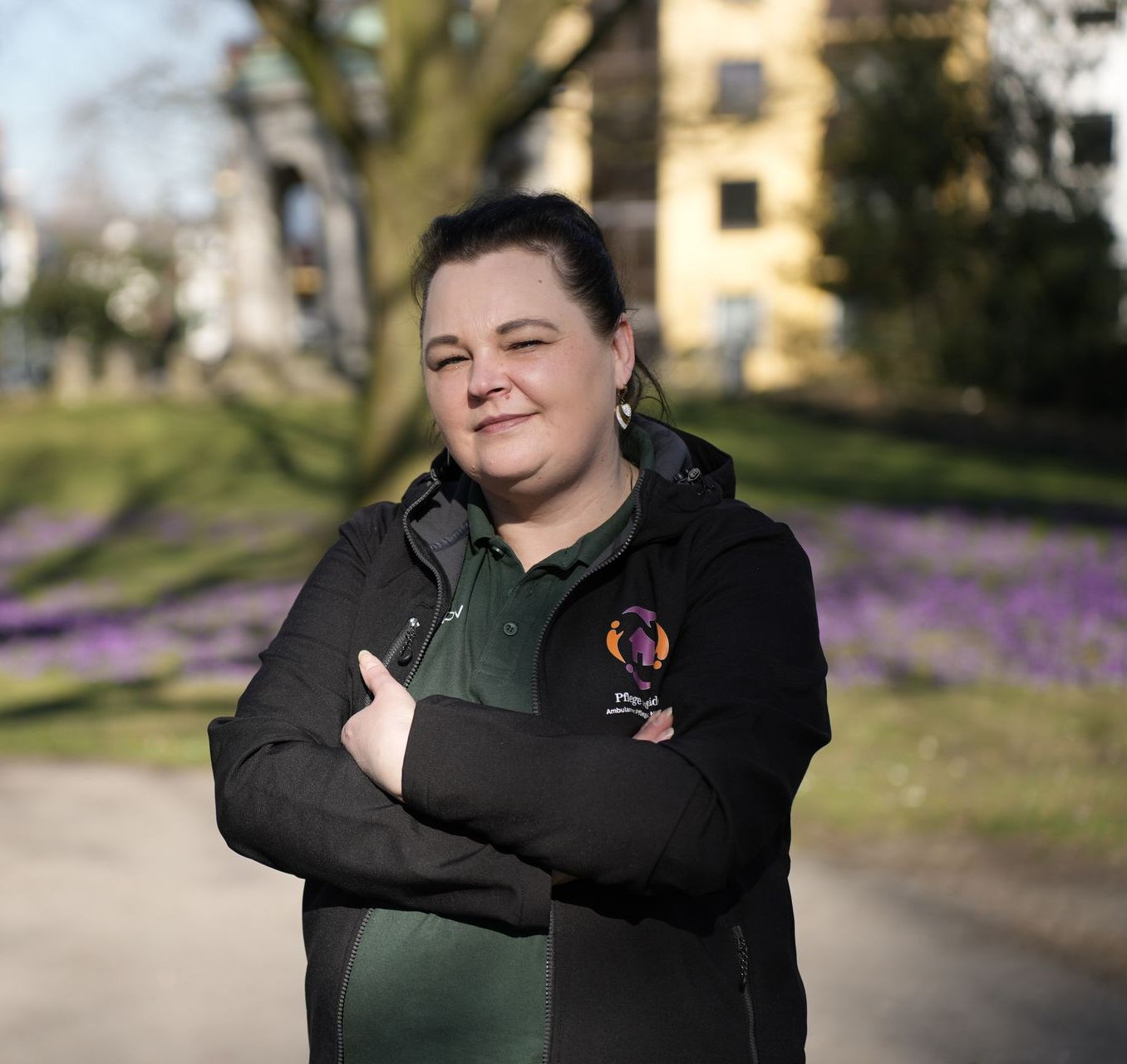 Eine Person mit dunklen Haaren trägt eine dunkle Jacke über einem grünen Hemd und steht mit verschränkten Armen in einem Park mit lila Blumen.