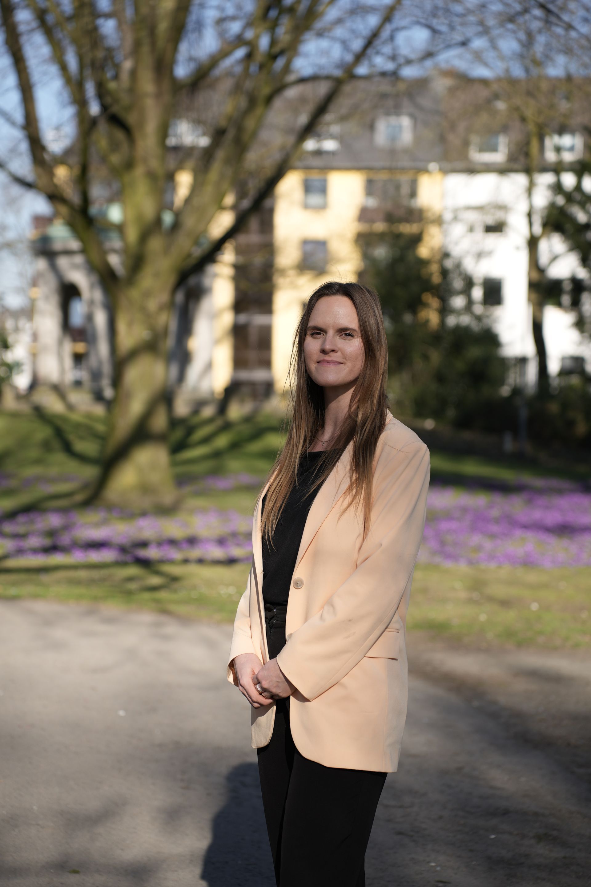 Eine Person in einem hellen Blazer steht im Freien vor einem Baum und einem Gebäude; im Hintergrund sind lila Blumen zu sehen.