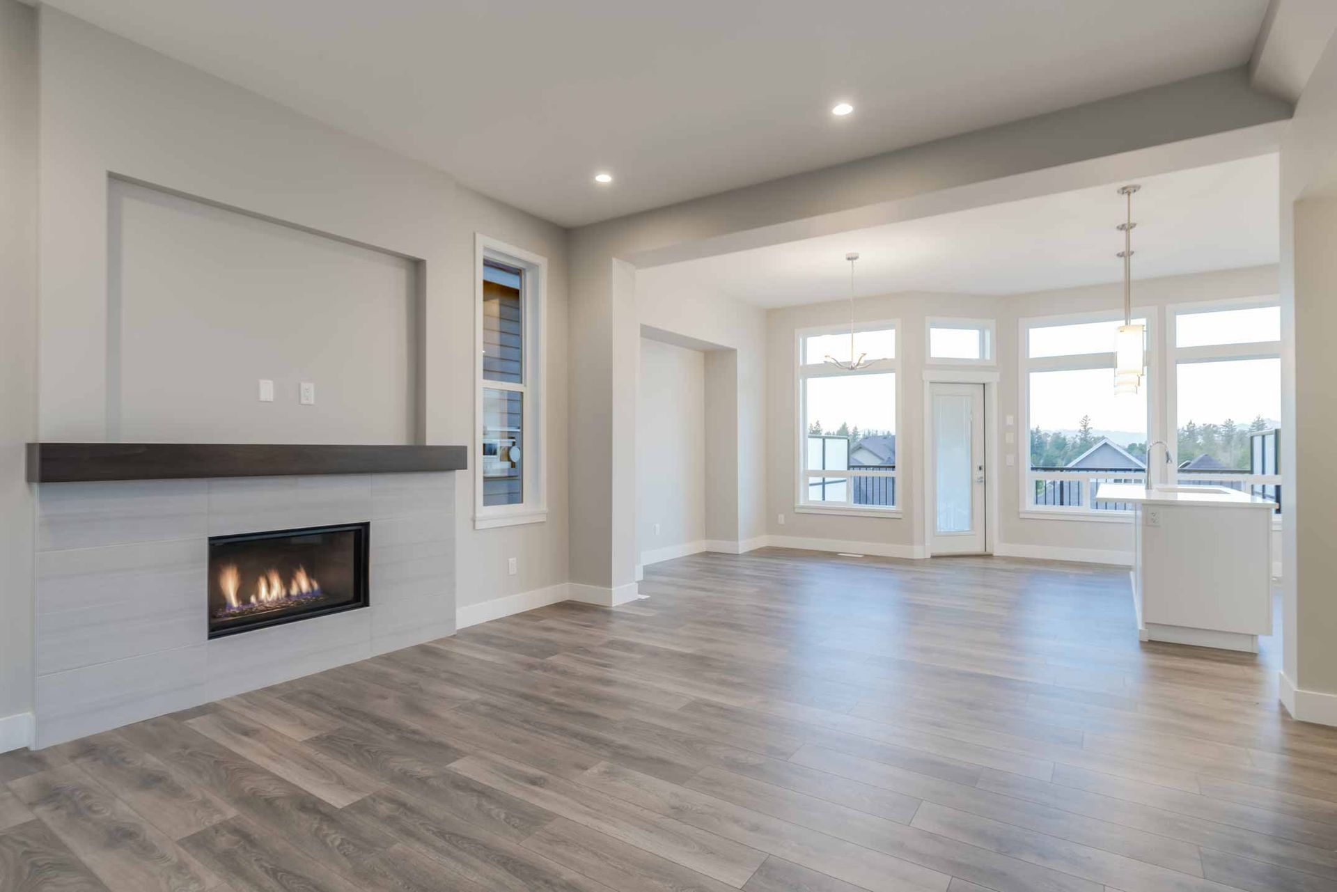 Modern open-concept living room with gray fireplace, wood floors, and large windows looking out to a deck