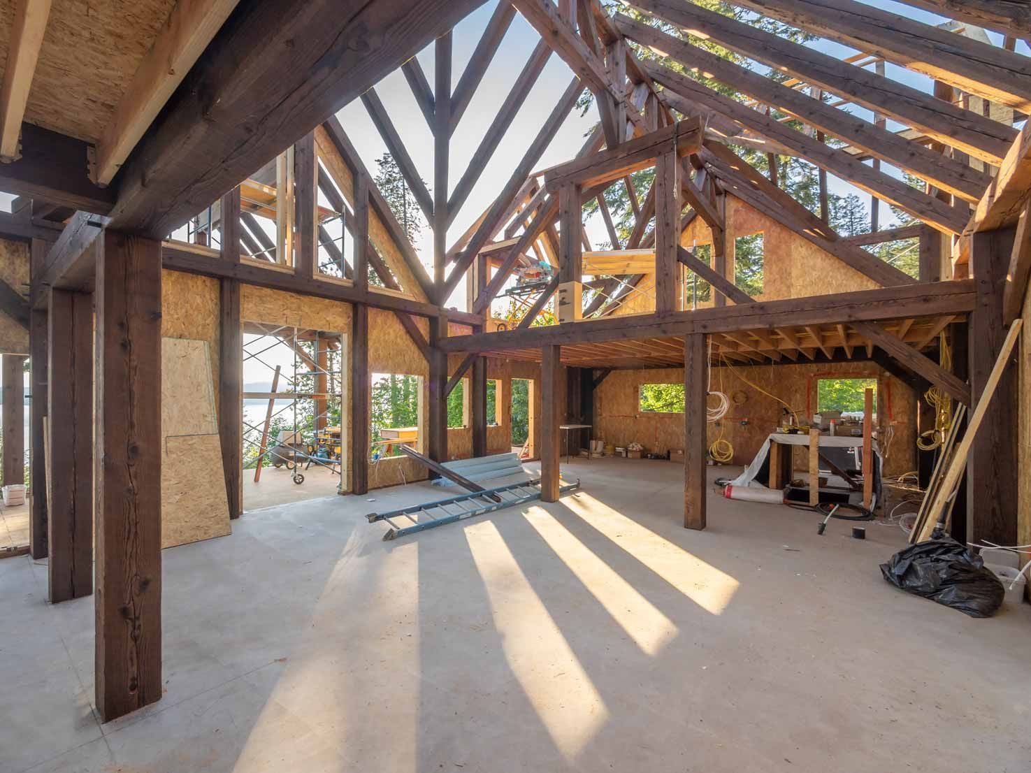 Unfinished timber-framed interior with exposed beams, large windows, and sunlight streaming across the floor