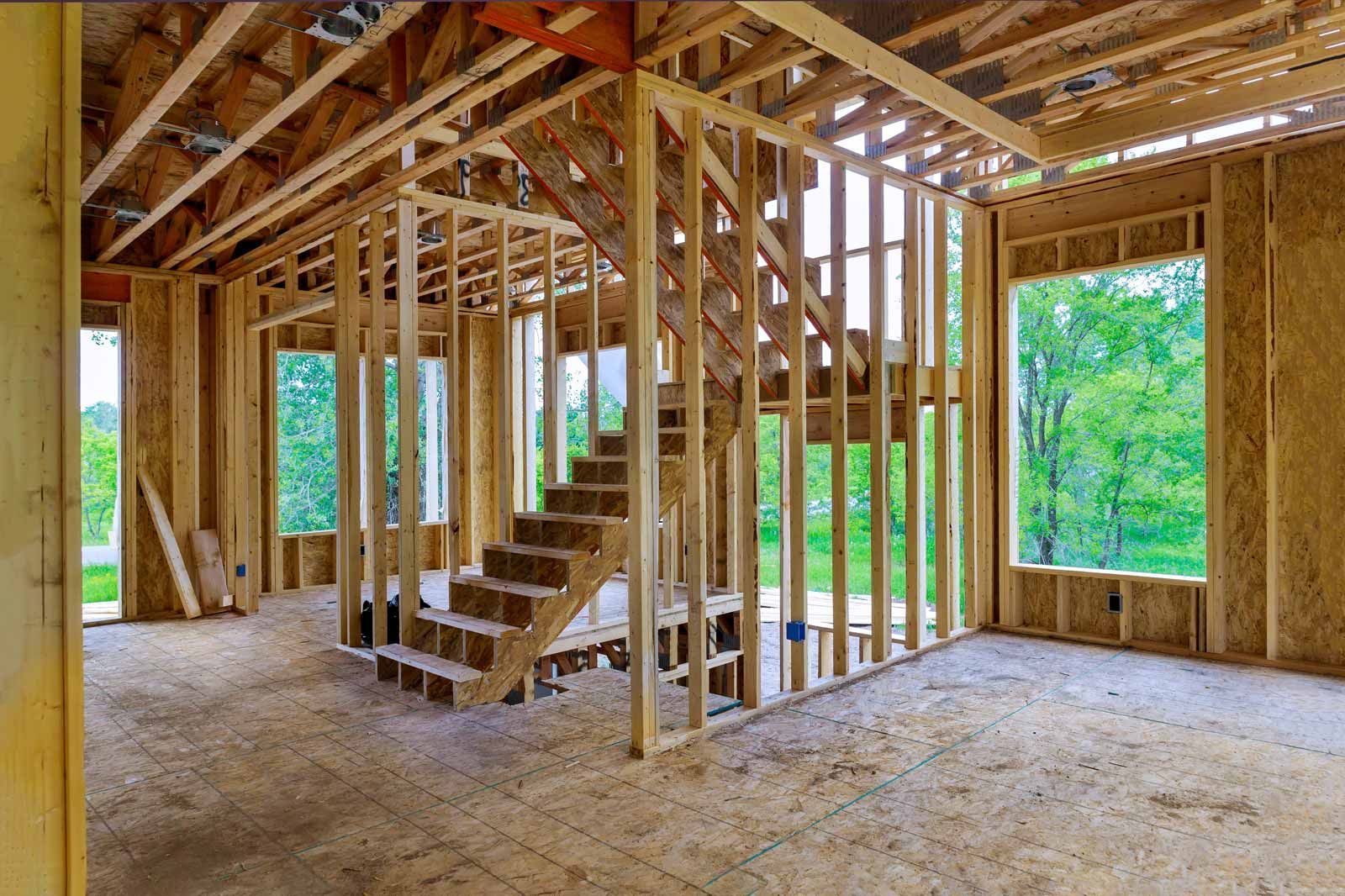 Unfinished house interior with exposed wooden framing, a staircase, and large windows overlooking greenery