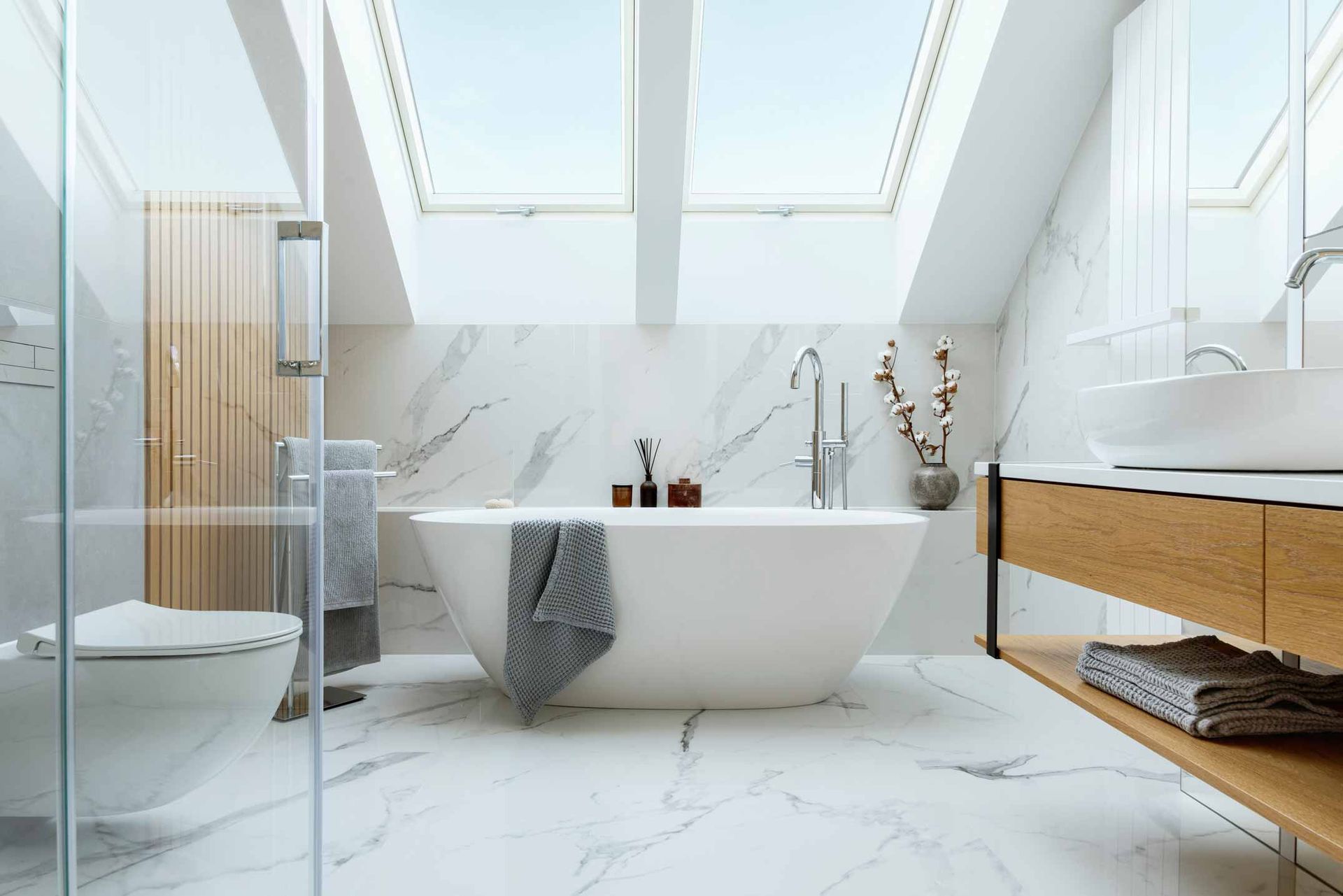 Bright modern bathroom with a freestanding tub, marble walls, skylights, and wooden vanity.