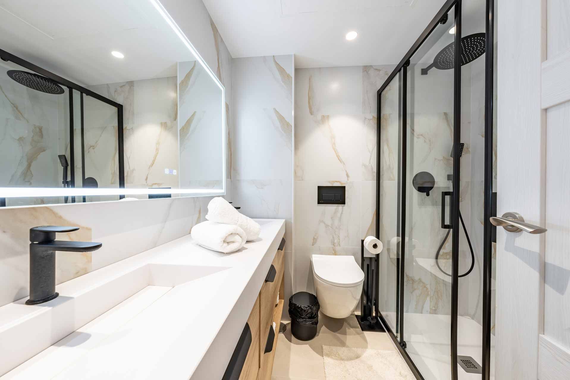 Modern bathroom with double vanity, large mirror, glass shower, and toilet in a bright, white interior