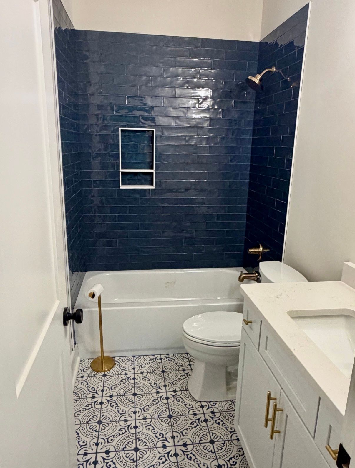 Small bathroom with dark blue tiled shower wall, white tub, toilet, and patterned floor tiles.