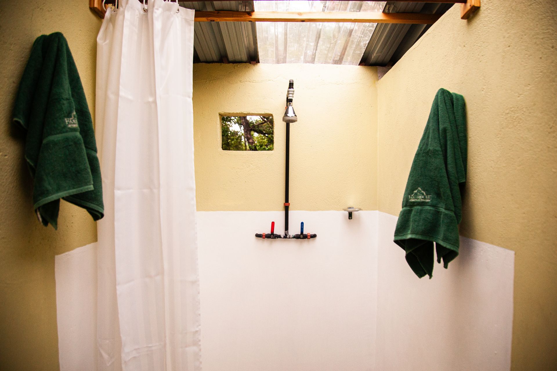 Outdoor shower with white walls, corrugated metal roof, green towels, and showerhead.