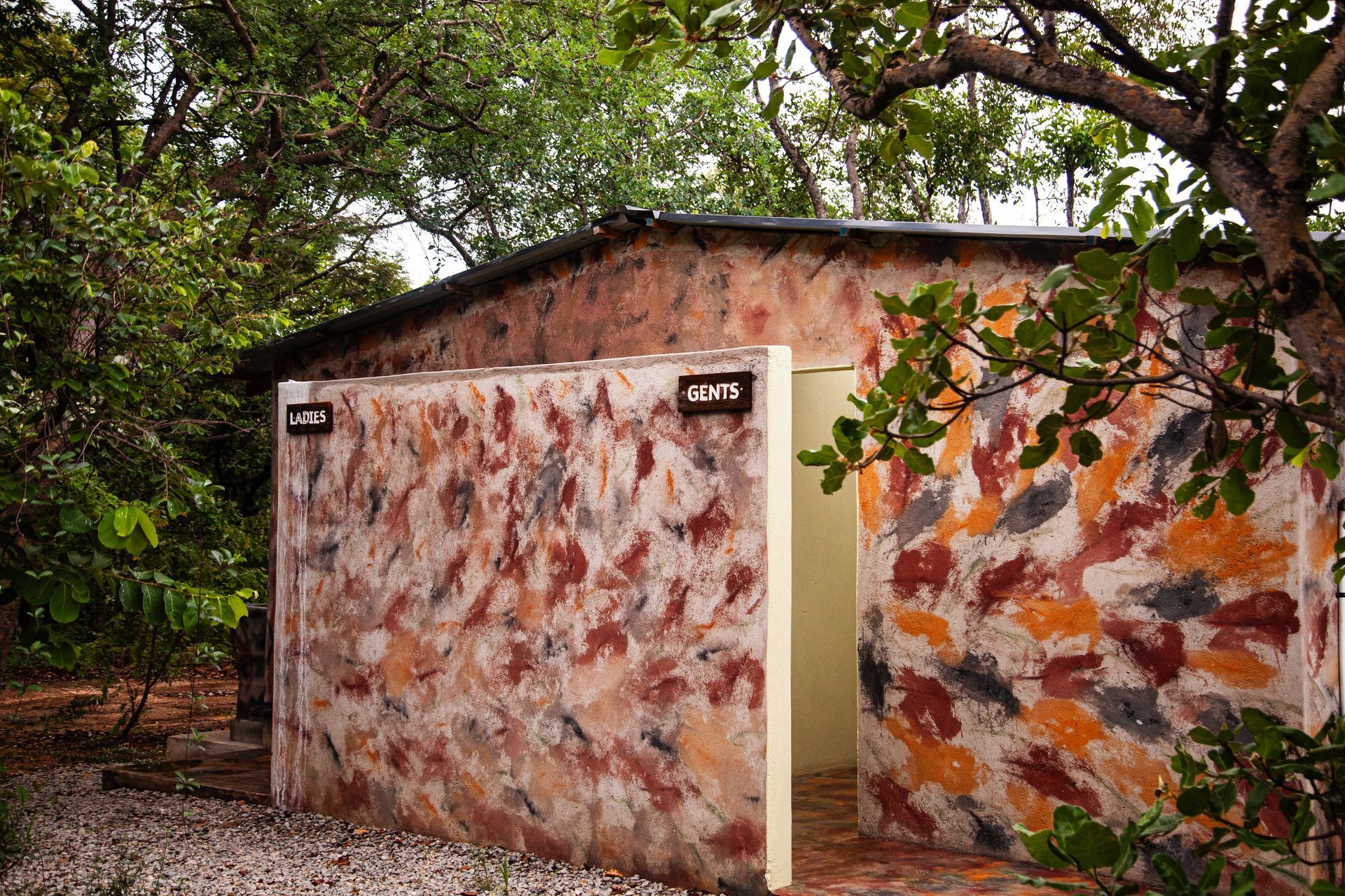 Small ablution building with open doorway, exterior walls covered in colorful mottled paint, surrounded by trees.