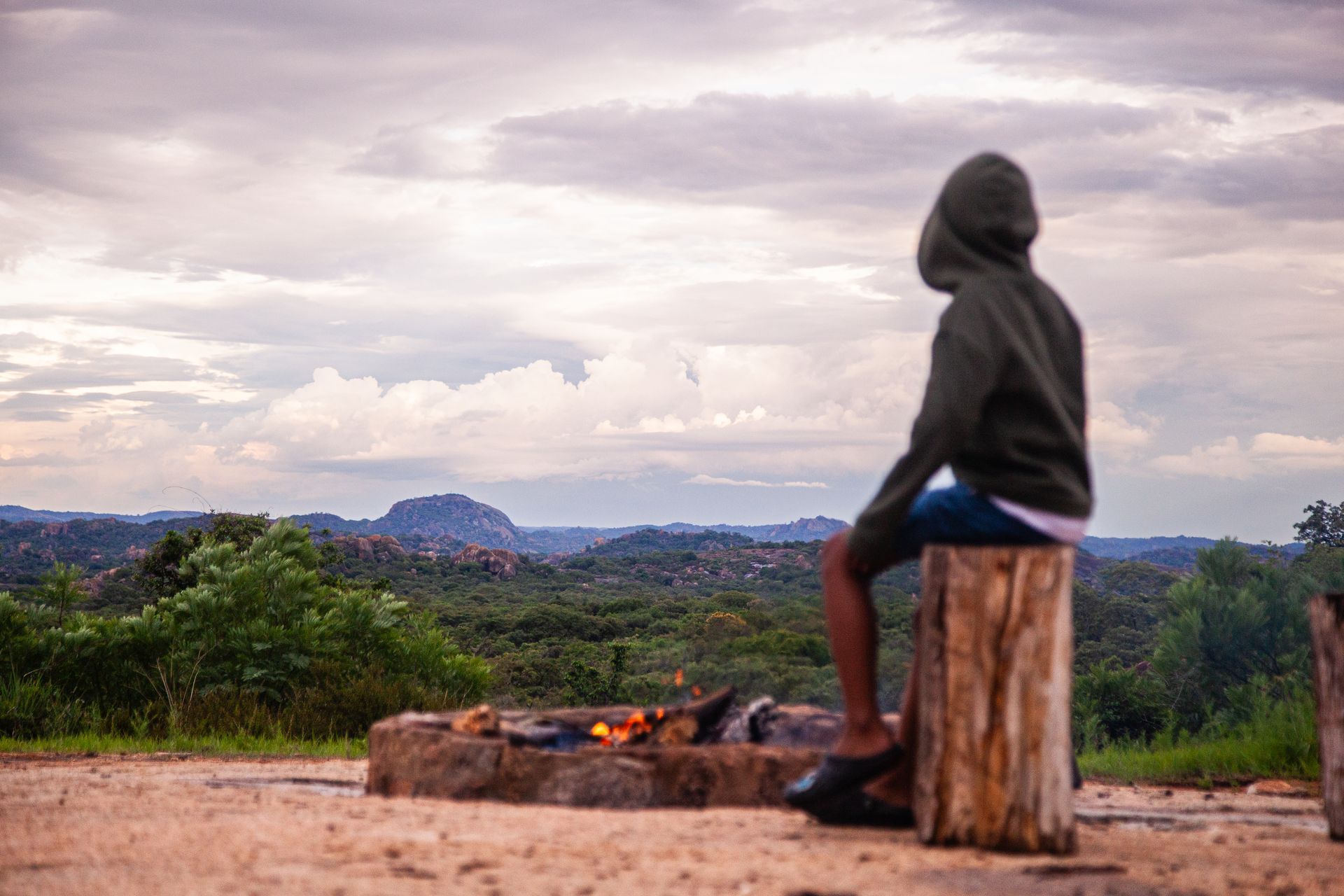 Person in a hoodie sits on a stump, gazing at a landscape with a fire pit in front. Overcast sky.