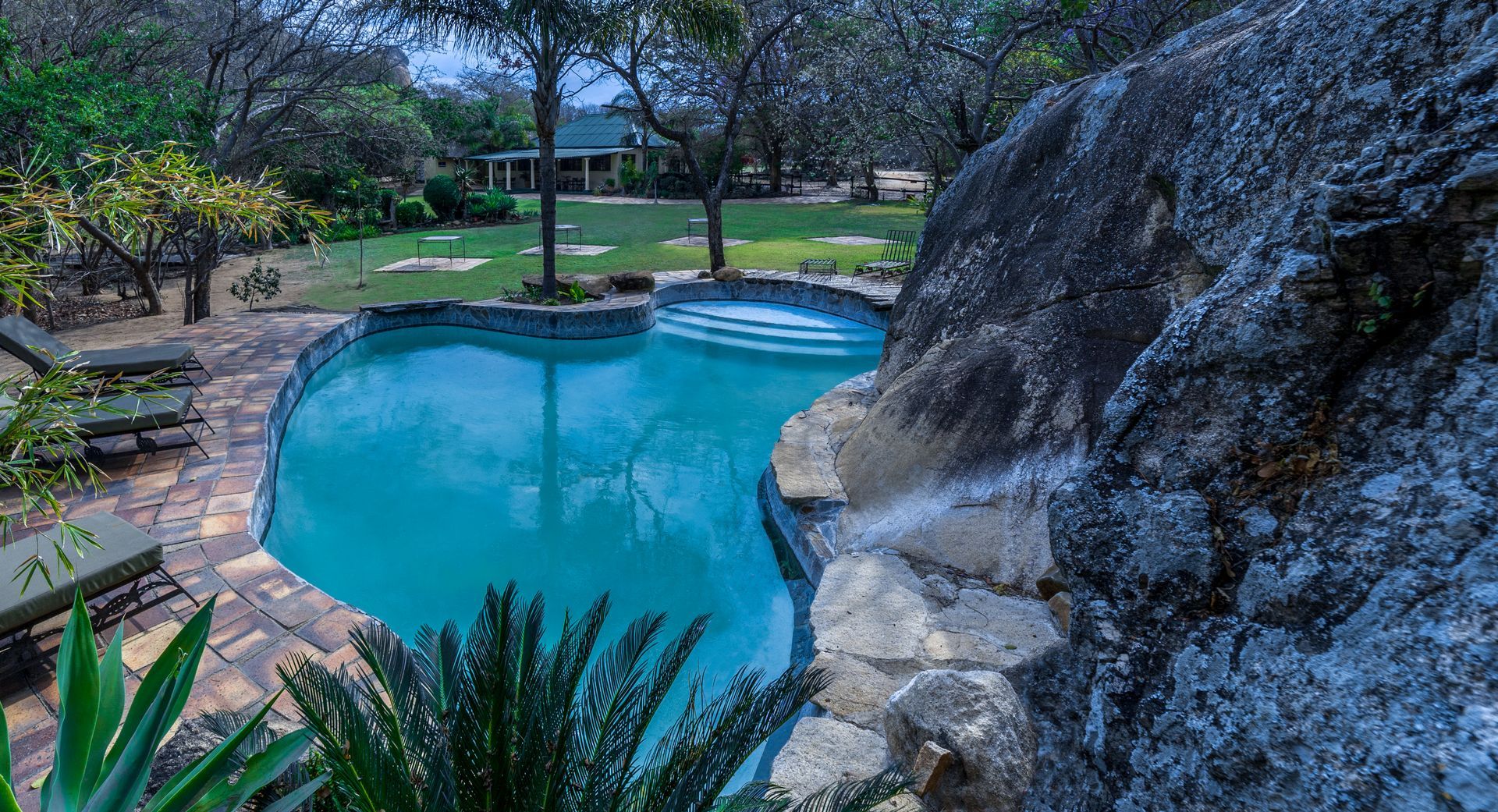 Pool with waterfall feature next to a rock face, surrounded by greenery and lawn.