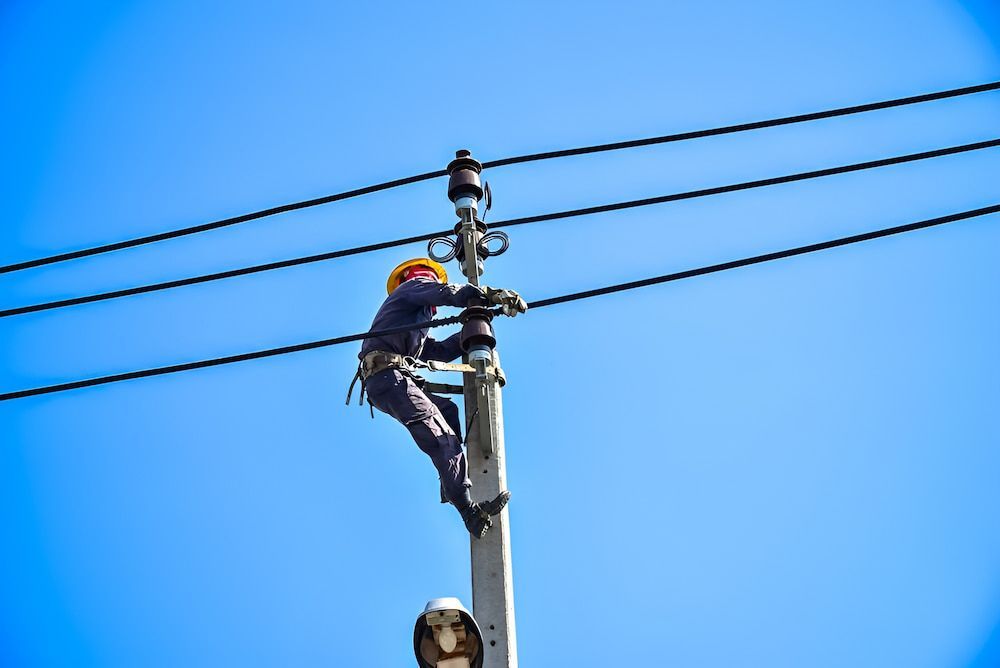 A Man Is Climbing A Power Pole With A Blue Sky — In-Line Electrical Southern Highlands in Moss Vale, NSW
