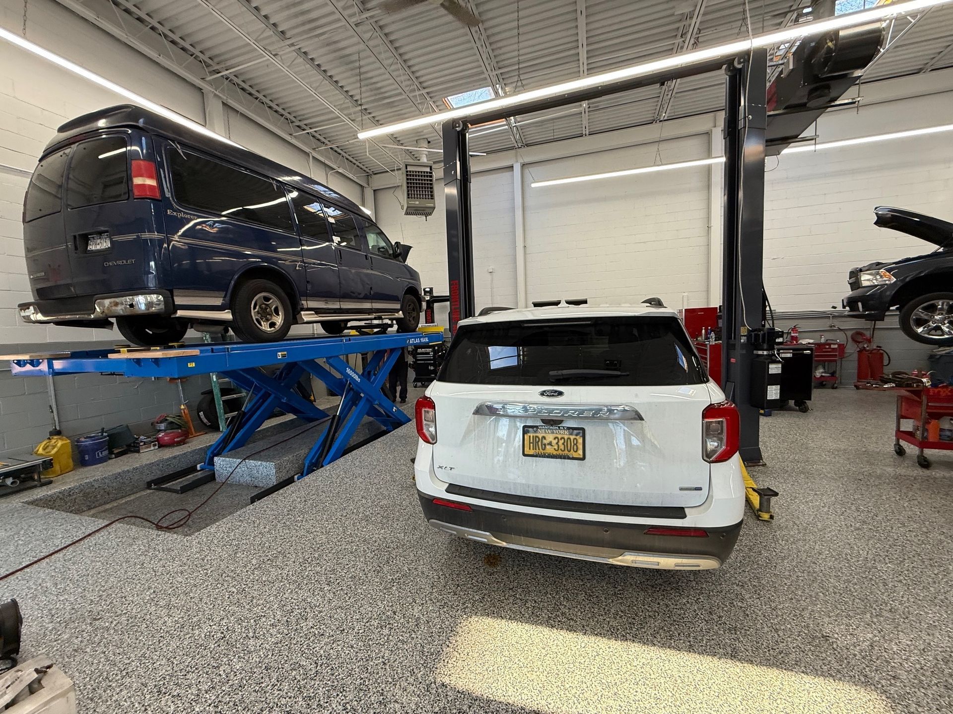 A modern auto repair shop with a dark blue van lifted on a scissor lift and a white SUV parked on the garage floor.
