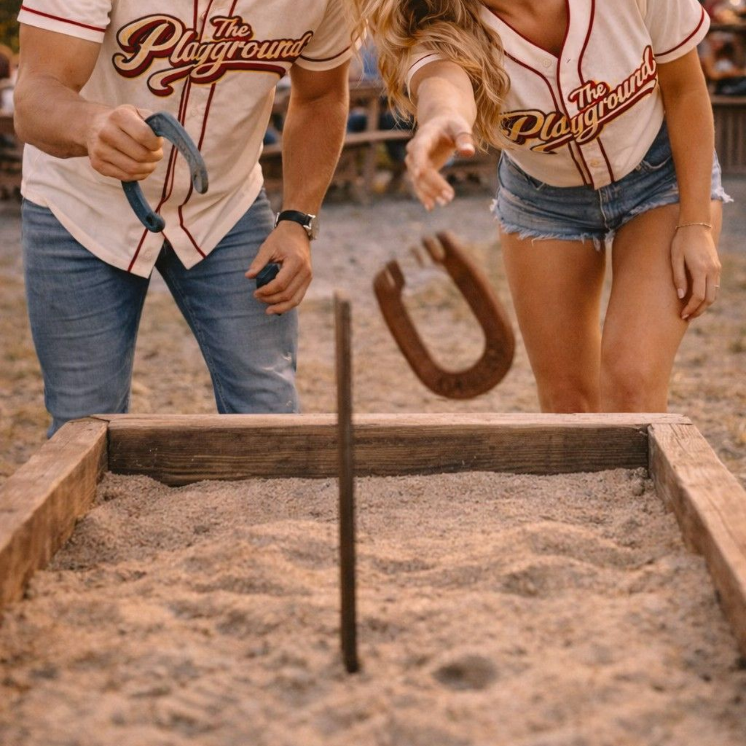 Two people in white shirts and denim shorts playing horseshoes in a sandy pit