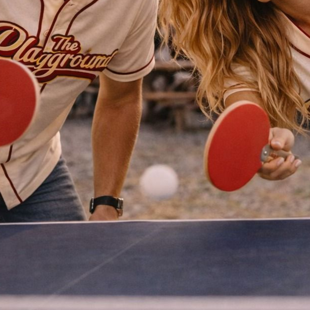 Two people playing ping pong, holding red paddles over a blue table outdoors.
