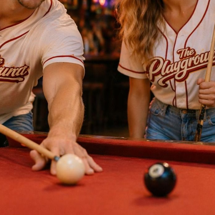 Two people lining up a pool shot at a red-felt table, with a white cue ball and black 8-ball visible.