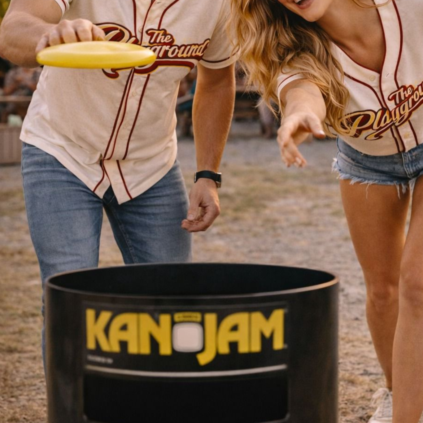 Two people playing KanJam outdoors, tossing a yellow flying disc toward a black target bin.