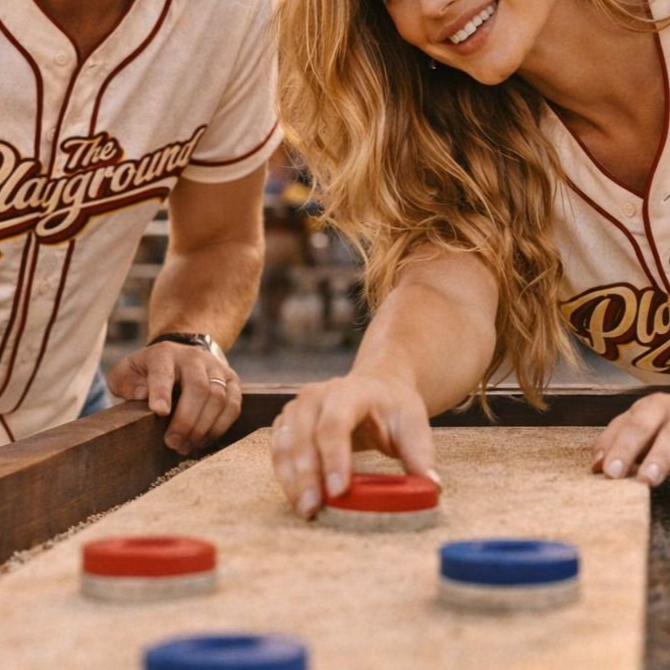 Two people playing tabletop shuffleboard, sliding a red puck toward blue and red pucks on a wooden table