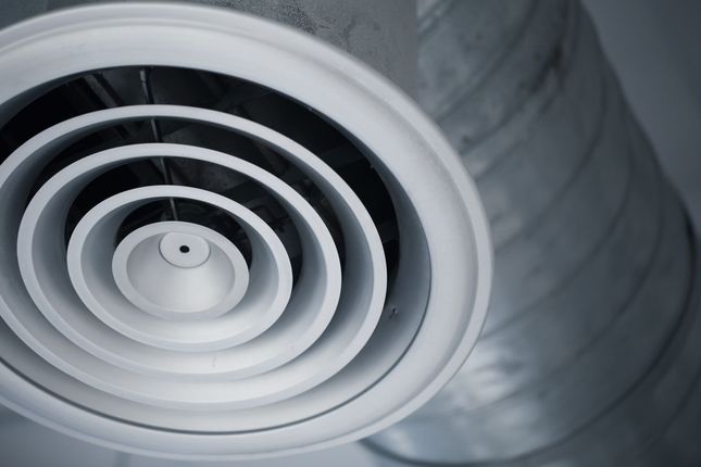 A white circular ceiling air diffuser vent connected to a silver industrial duct.